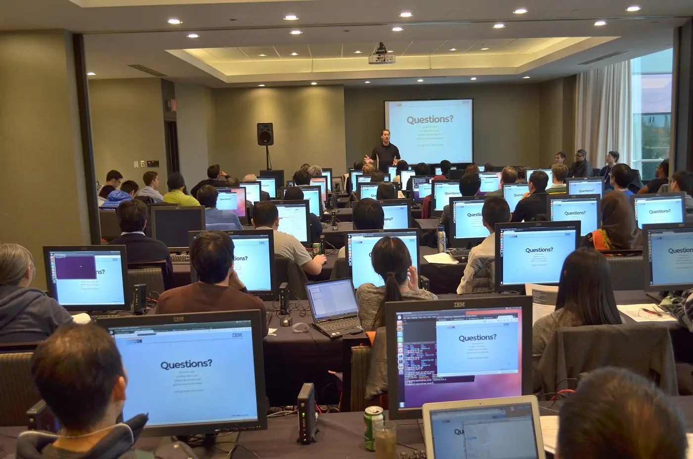 Salle de formation informatique : une trentaine de participants devant des ordinateurs portables, face à un écran de présentation.