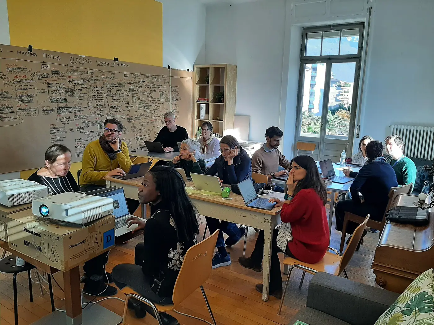 Atelier collaboratif : plusieurs personnes travaillent autour de tables avec des ordinateurs portables, devant des murs couverts de notes.